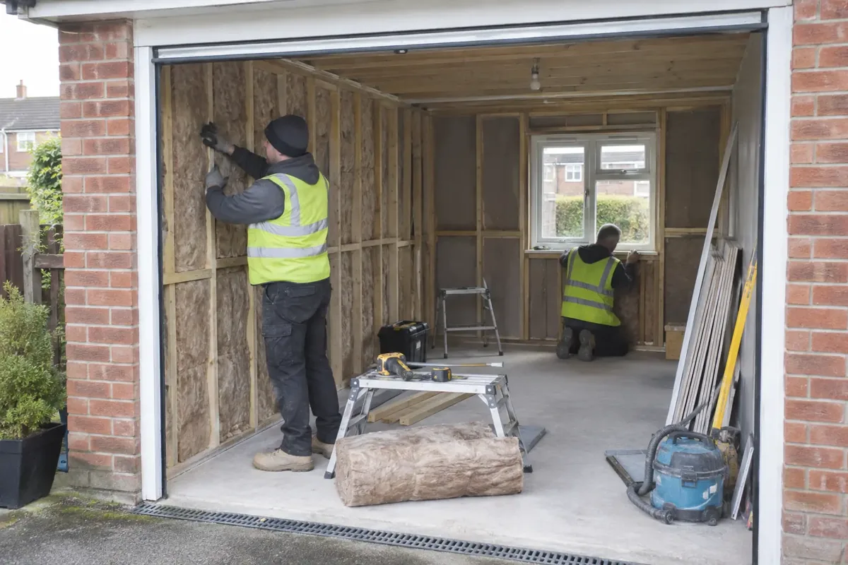 Builders converting an attached brick garage into a habitable room, new insulated internal stud lining and small installed front window visible within original wide garage opening, worker fitting insulation and checking frame, appropriate PPE and tidy site.